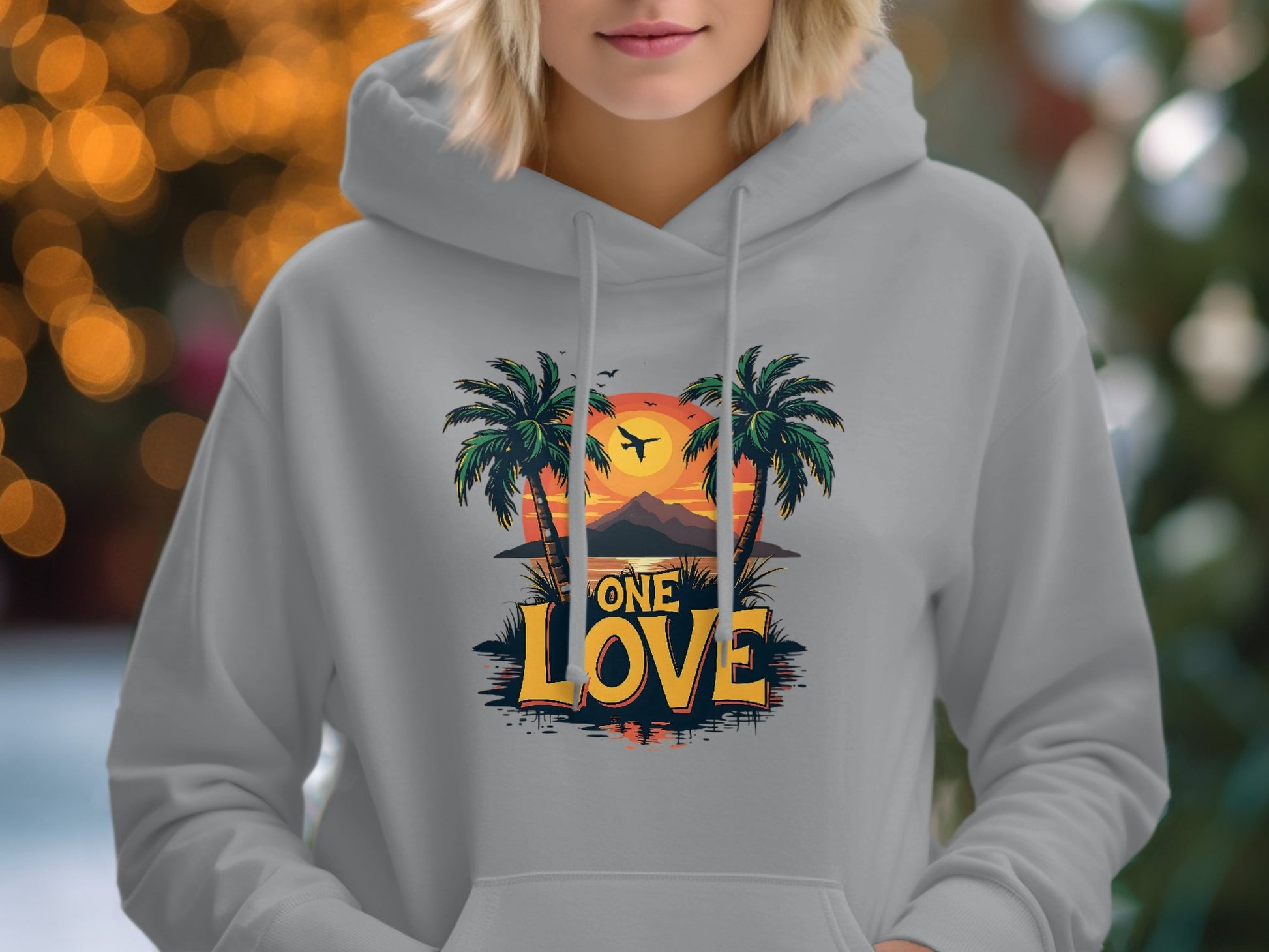 A person wears a Garment Graphics light gray hoodie featuring a unique tropical design of two palm trees against a sunset and the phrase One Love, with a softly blurred background aglow with warm, bokeh lights enhancing its charm.