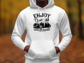 A person wearing a classic fit white hoodie featuring ENJOY Classic Car and a vintage car graphic exudes the community spirit of the Vintage Ride Community Aircooled. The autumn trees in the blurred background enhance the scene. (Brand: Garment Graphics).