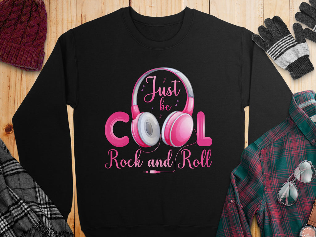 The Garment Graphics black sweatshirt showcases a pink and white Just be cool, Rock and Roll Headphones design. Perfect for music fans, its styled next to a red plaid shirt, hat, scarf, and gloves on wood. An ideal choice for fashion lovers seeking rhythm-infused style.