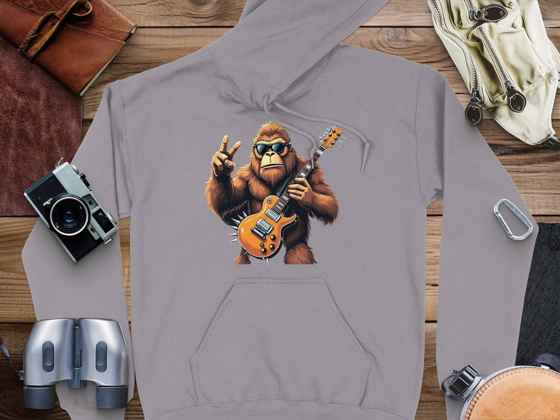 Garment Graphics presents a gray gorilla hoodie with an ape in sunglasses, playing an electric guitar and making a peace sign. Perfect for music lovers, it is styled on a wooden surface with accessories including a camera, binoculars, and leather pouch.