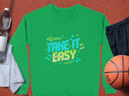 Garment Graphics unisex green sweatshirt, featuring Take It Easy in colorful letters with musical notes and arrows, lays comfortably on a sports surface alongside a water bottle, gray towel, black shorts, and an orange basketball.