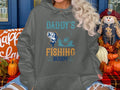 A person in a Garment Graphics hoodie featuring Daddys Fishing Buddy with a fish and man in a boat sits amid autumn decor like pumpkins, leaves, and a scarecrow—ideal for outdoor adventure enthusiasts.