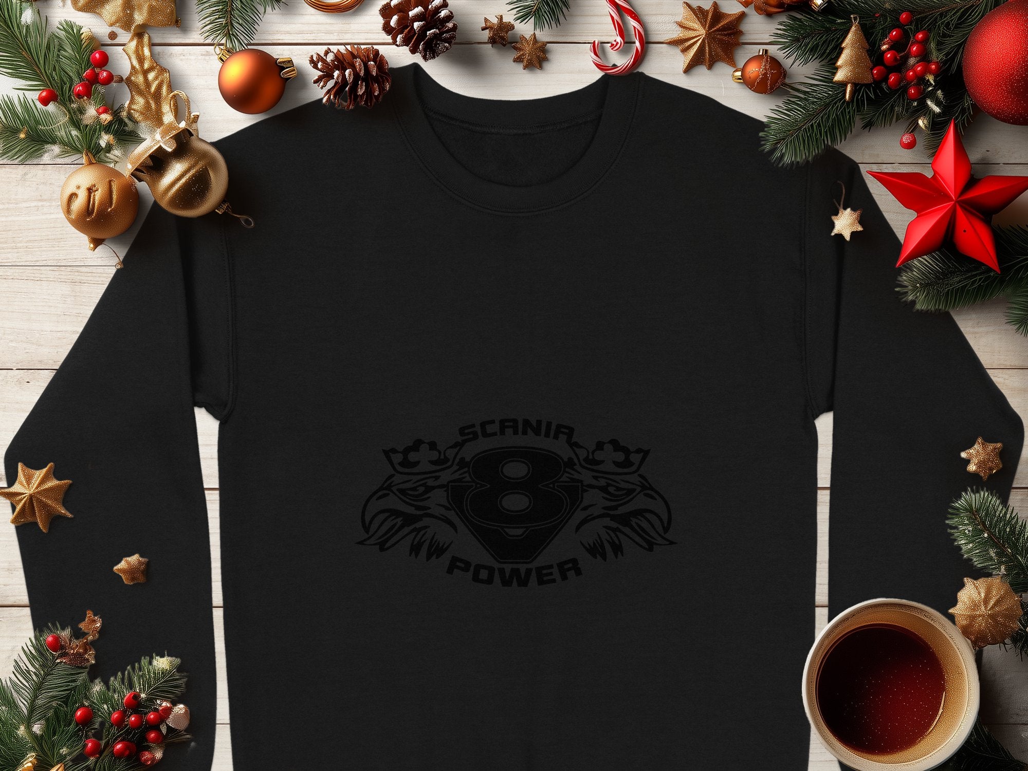 A stylish black long-sleeve shirt by Garment Graphics showcasing the Scania V8 Power design is artfully displayed with Christmas decorations such as pine cones, ornaments, stars, and a cozy coffee cup on a wooden surface, highlighting its cool print.