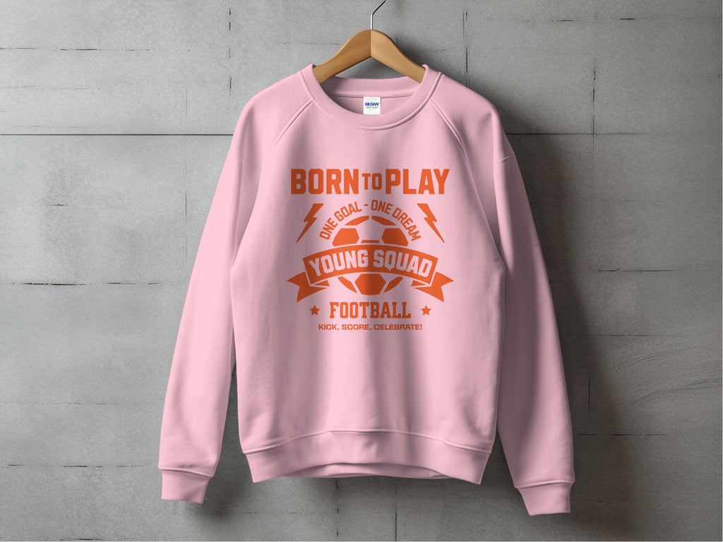 A young footballers pink sweatshirt by Garment Graphics hangs on a concrete wall, featuring an orange graphic with BORN TO PLAY, YOUNG SQUAD FOOTBALL, and KICK. SCORE. CELEBRATE. Includes a football, goalpost, and banners. Available in ideal unisex sizing.