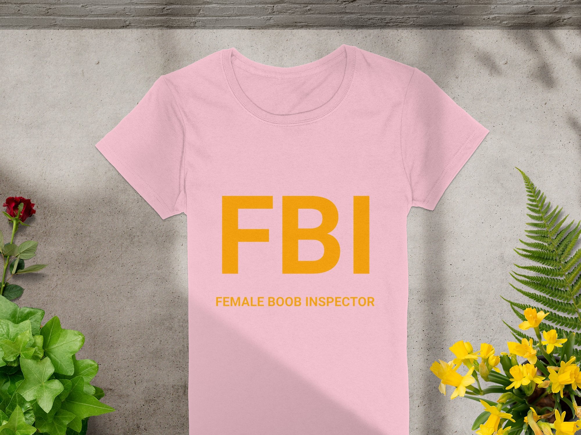 A hilarious gag gift, the Garment Graphics FBI Female Boob Inspector T-shirt for women features vibrant yellow text on a pink fabric, showcased on a concrete surface surrounded by lush green plants and cheerful yellow flowers for a playful vibe.