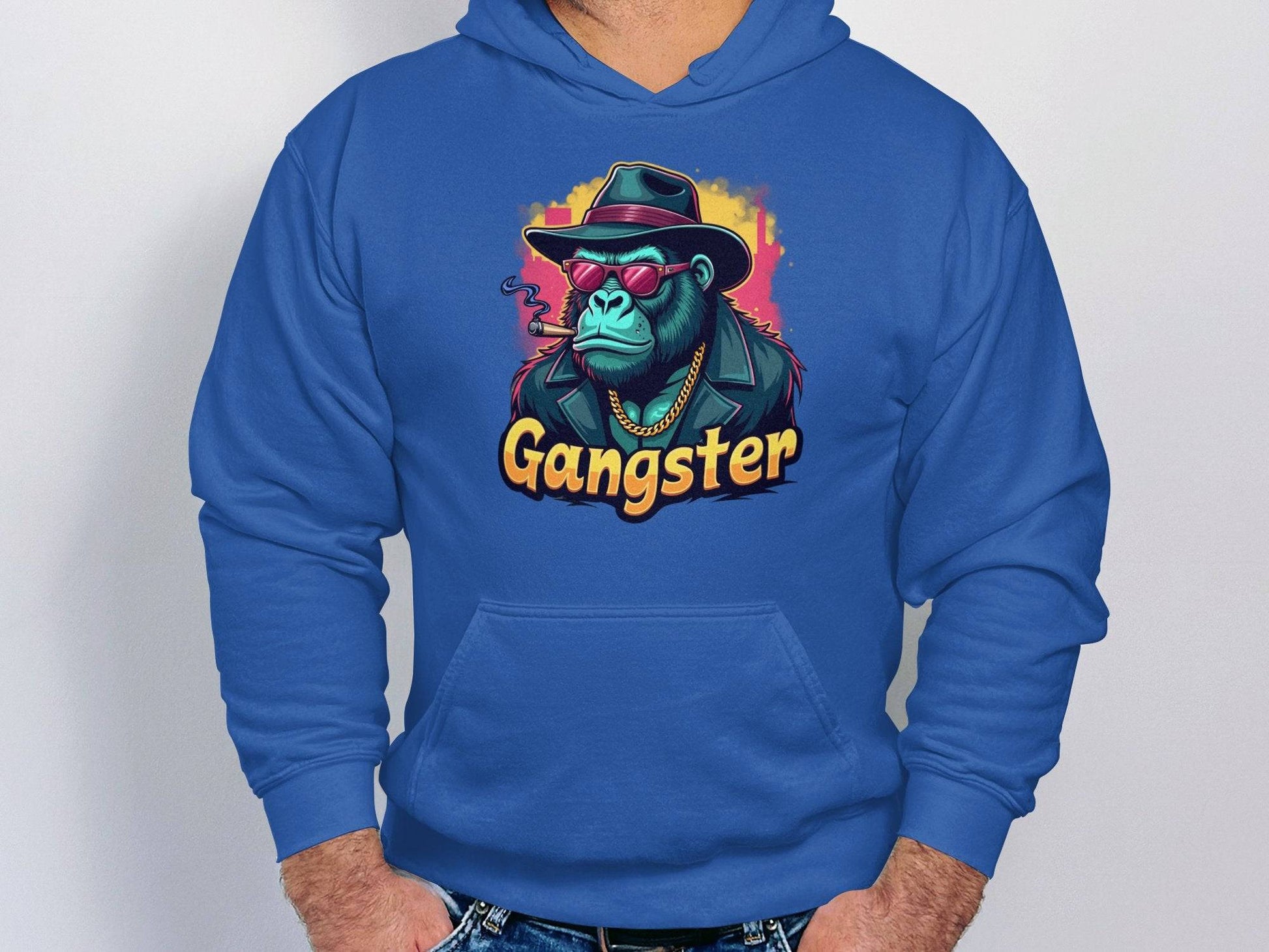 A person wearing a Garment Graphics blue hoodie, featuring a gangster gorilla with a hat, sunglasses, and cigar, exudes smokey cool. The word Gangster is printed below the graphic on the medium-heavy fabric as they stand against a plain backdrop with hands in pockets.