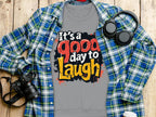 A Garment Graphics gray t-shirt with Its a Good Day to Laugh in bold text rests on a wooden surface. Perfect for unisex sizing, its complemented by a blue plaid shirt, headphones, a camera, and a cup—a cozy and stylish ensemble ready for any adventure.