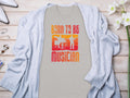 A gray cardigan by Garment Graphics is displayed flat lay style on wood, with a beige Born To Be Musician T-shirt underneath, showcasing drummer and guitarist silhouettes. Nearby accessories and white flowers complete the look, ideal for music lovers.