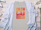 A gray cardigan by Garment Graphics is displayed flat lay style on wood, with a beige Born To Be Musician T-shirt underneath, showcasing drummer and guitarist silhouettes. Nearby accessories and white flowers complete the look, ideal for music lovers.