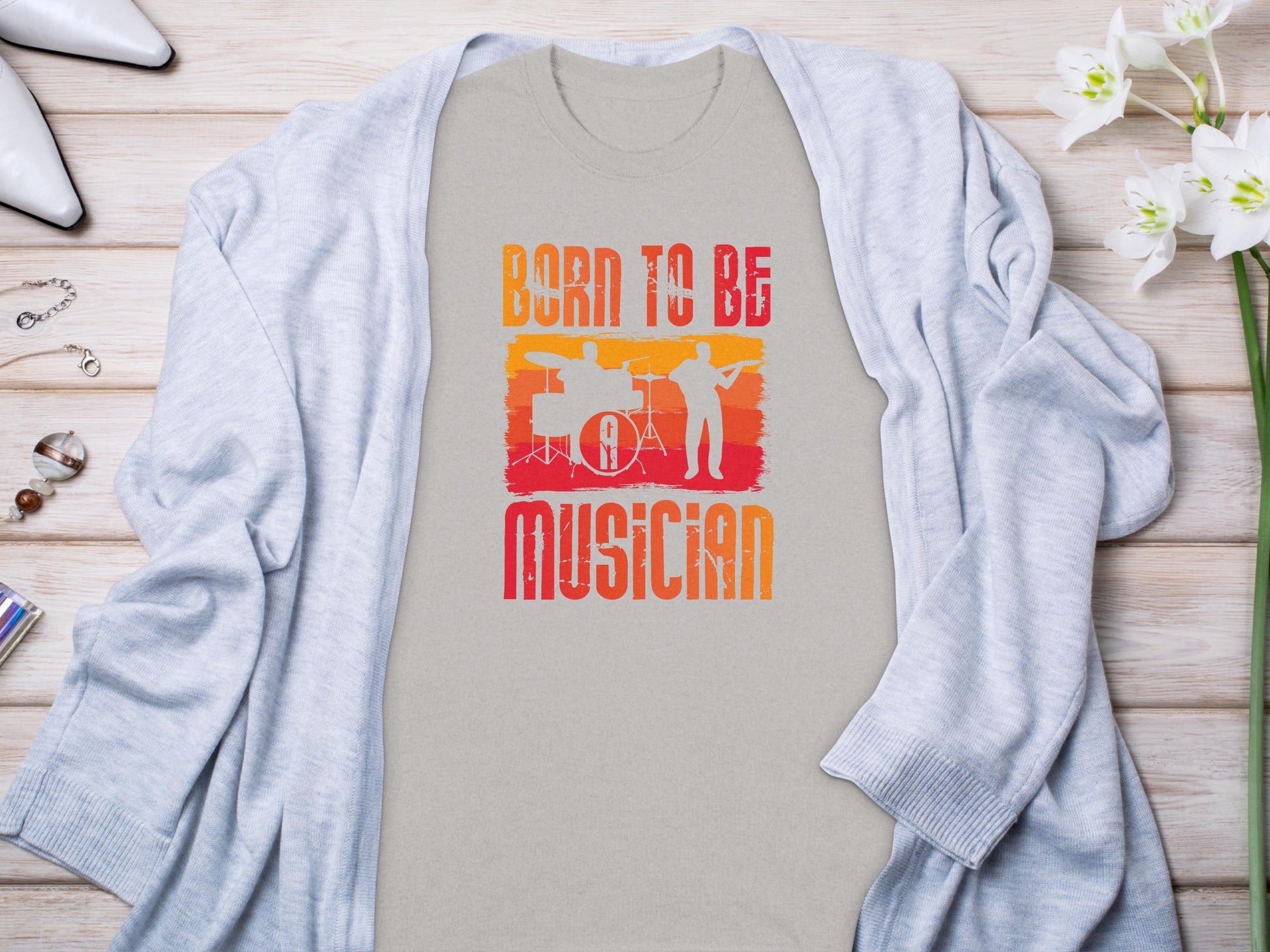 A gray cardigan by Garment Graphics is displayed flat lay style on wood, with a beige Born To Be Musician T-shirt underneath, showcasing drummer and guitarist silhouettes. Nearby accessories and white flowers complete the look, ideal for music lovers.