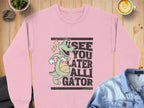 Garment Graphics pink unisex sweatshirt features a cartoon alligator holding a basketball with the phrase See You Later Alligator. Its displayed on a wooden surface next to a denim jacket, potted plant, and cappuccino.