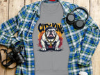 A gray shirt from Garment Graphics Bulldog Sweatshirts, featuring a bulldog with a gold chain and City King cap, lies on wood beside a blue/green plaid shirt, headphones, camera, and white travel mug.