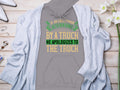 The Garment Graphics gray hoodie displays a witty quote: If an Englishman gets run over by a truck, he apologizes to the truck in vivid yellow and green. It is artfully laid flat on a white surface with a light gray cardigan, white flowers, and assorted accessories.