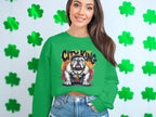 A long-haired woman stands against a shamrock backdrop wearing a green Garment Graphics Bulldog Sweatshirt, featuring a gold chain, bulldog illustration, and City King text, paired with light blue denim shorts.