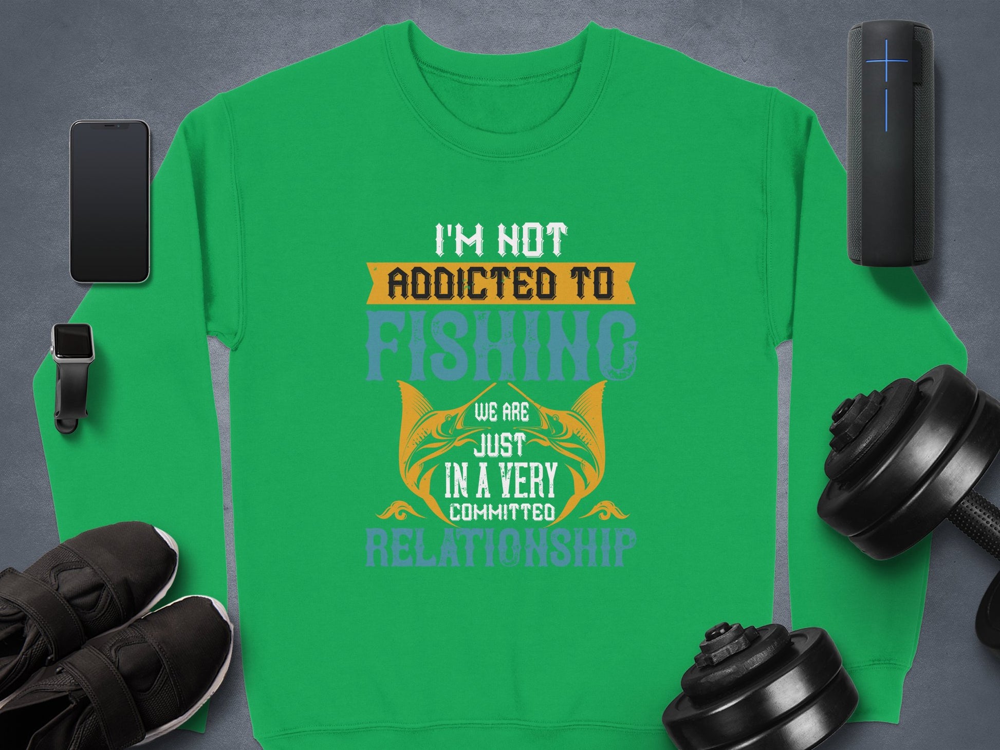 Garment Graphics green sweatshirt with the fun text Im Not Addicted to Fishing, We Are Just in a Very Committed Relationship in different fonts/colors. Unisex sized, its displayed on a gray backdrop surrounded by a smartphone, earbuds, wireless speaker, black shoes, and dumbbells.