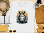 On a wooden surface lies a white Garment Graphics sweatshirt with an illustrated owl design. Surronding it are a scarf, quilted jacket, sunglasses, mug, and notebook with pen in yellow and brown hues. The unisex sizing makes this cozy ensemble perfect for everyone.