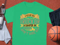A green Garment Graphics t-shirt says: If hes got golf clubs in his truck or a camper in his driveway, I dont hire him. Perfect for camping humor fans, its artistically displayed on a brown textured background with a water bottle, shorts, towel, and basketball nearby.