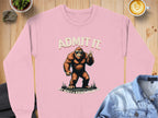 Garment Graphics pink Admit It Sweatshirt showcases a Bigfoot graphic holding two fingers. The text reads ADMIT IT above and LIFE WOULD BE BORING WITHOUT ME below. Its displayed on wood, accompanied by coffee and a denim jacket.