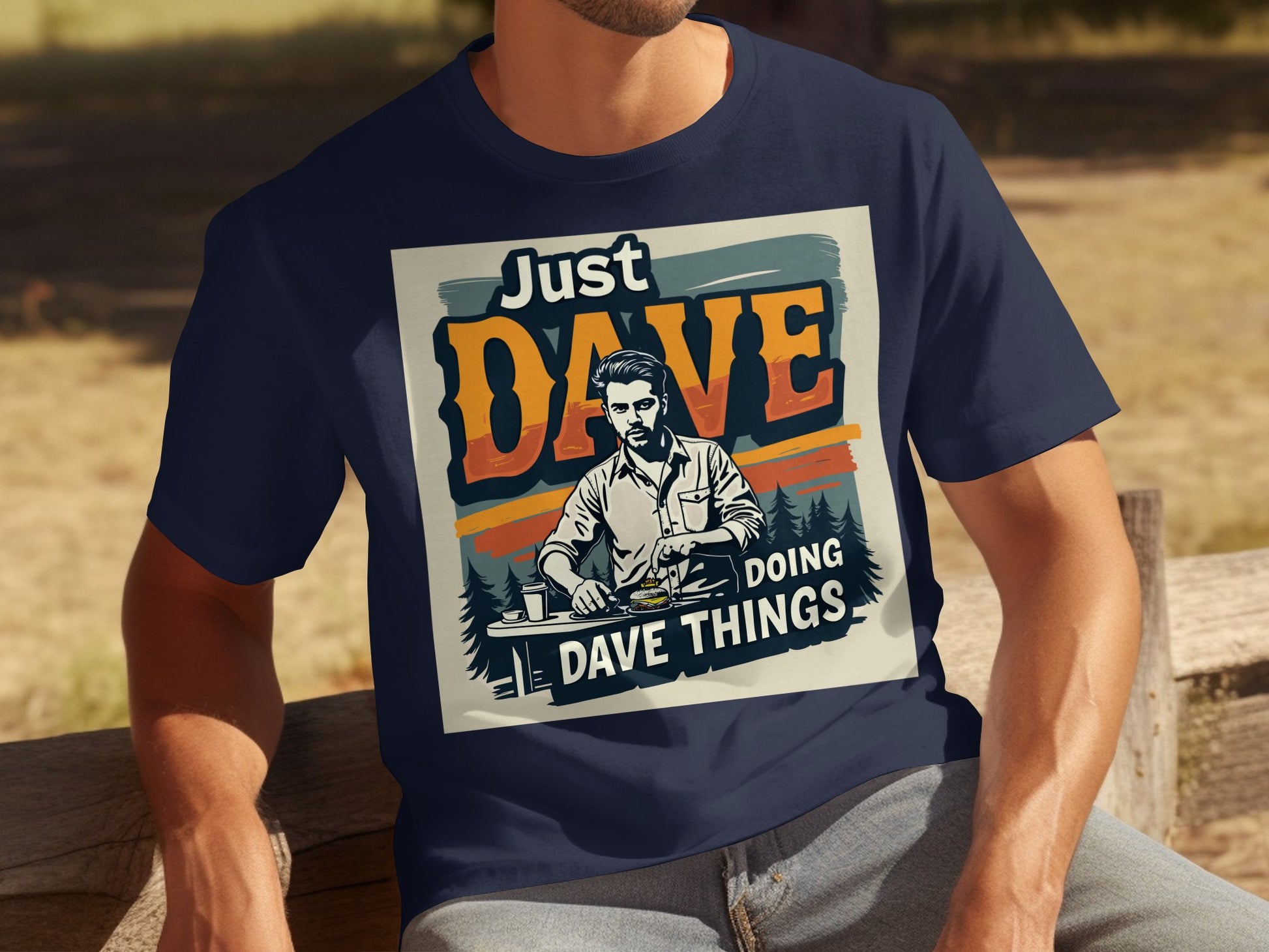 A person sits on a wooden bench wearing a classic navy blue T-shirt by Garment Graphics. The vintage-style shirt features the words Just Dave Doing Dave Things with an image of a man in a serene forest setting.