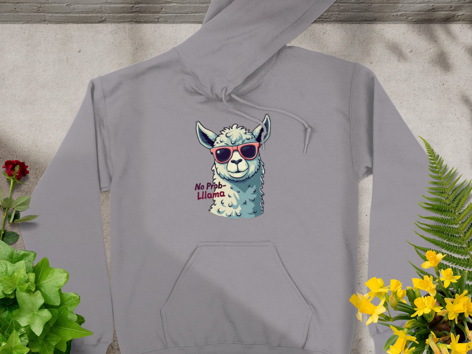 A gray hoodie from Garment Graphics, featuring a llama in red sunglasses and the text No Prob-llama, sits on a concrete surface. Its framed by greenery and yellow flowers.