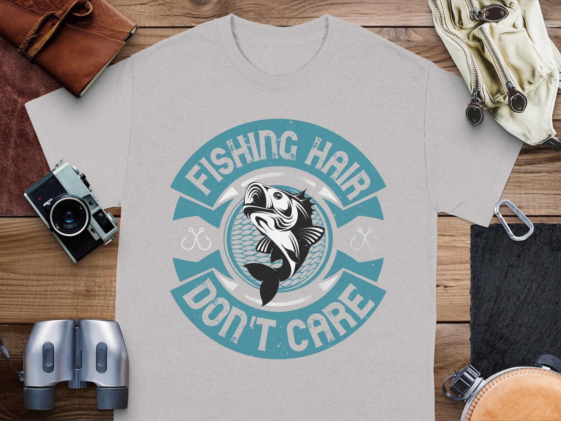 A Garment Graphics gray t-shirt with a unique fishing design, featuring a stylized fish and Fishing Hair Dont Care in blue and white, is ideal for fishing enthusiasts. It’s displayed on wood, accompanied by a camera, binoculars, and gloves.