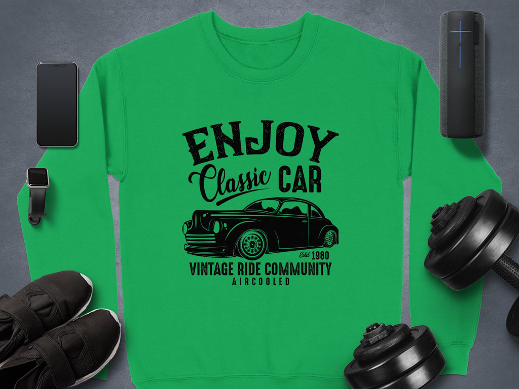 A Garment Graphics green sweatshirt with a black vintage car graphic, ENJOY Classic Car, and Vintage Ride Community Aircooled Estd 1980. Nearby are a smartphone, smartwatch, Bluetooth speaker, sneakers, and dumbbells.
