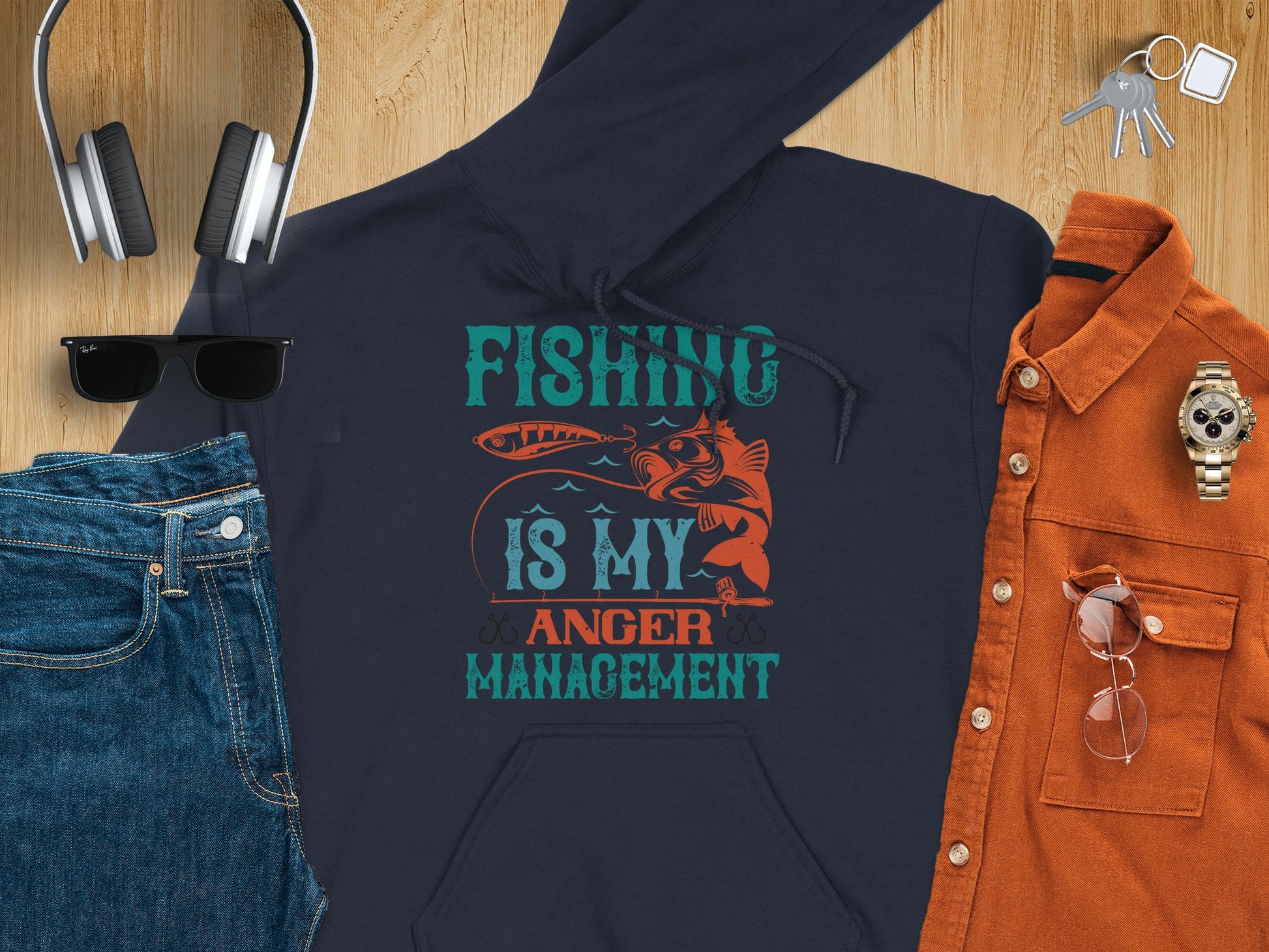 A flat lay reveals an anglers outfit featuring a Garment Graphics hoodie with Fishing is my anger management text, complemented by headphones, keys, denim jeans, sunglasses, a watch, and an orange shirt with glasses.