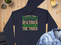 On a wooden table, a navy blue hoodie by Garment Graphics humorously reads: If an Englishman gets run down by a truck, he apologizes to the truck. A denim jacket, latte with leaf art, and small plants accompany this charming blend of fashion and English etiquette.