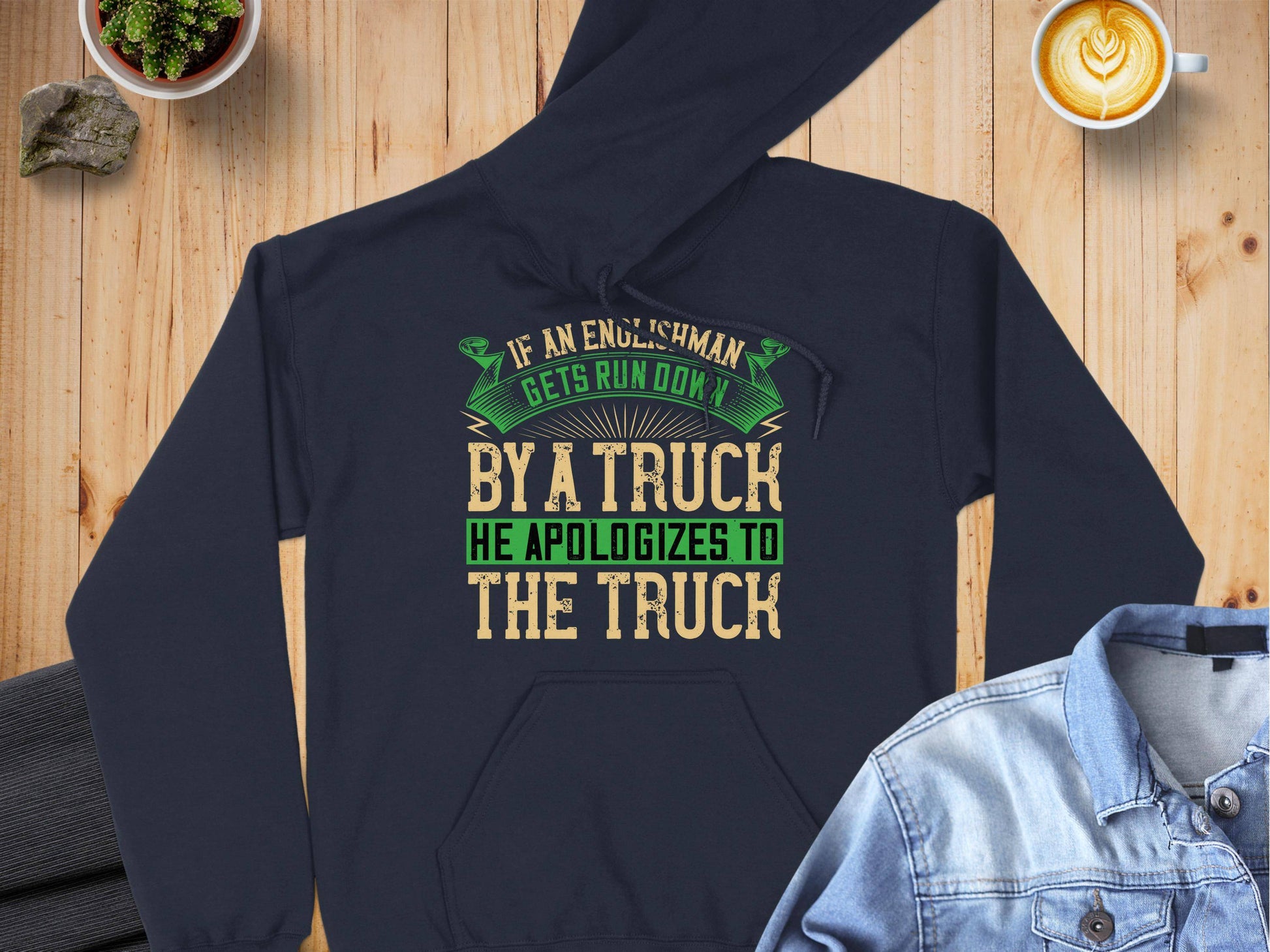 On a wooden table, a navy blue hoodie by Garment Graphics humorously reads: If an Englishman gets run down by a truck, he apologizes to the truck. A denim jacket, latte with leaf art, and small plants accompany this charming blend of fashion and English etiquette.