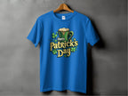 A blue statement T-shirt by Garment Graphics hangs on a wooden hanger against a gray background, featuring a green beer mug, shamrocks, and bold yellow and green text Happy Patricks Day, perfect for festive celebrations.