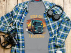 A Garment Graphics gray t-shirt with a bold truck design and KEEP ON TRUCKING text is on a wooden surface, surrounded artfully by a blue and green plaid shirt, headphones, camera, and white tumbler.