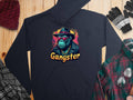 The Garment Graphics black hoodie, illustrating a gangster gorilla with sunglasses, hat, and chain above Gangster, is surrounded by a beanie, gloves, plaid shirt, and scarf on a wooden surface. Made from medium-heavy fabric for ultimate comfort in smokey cool style.