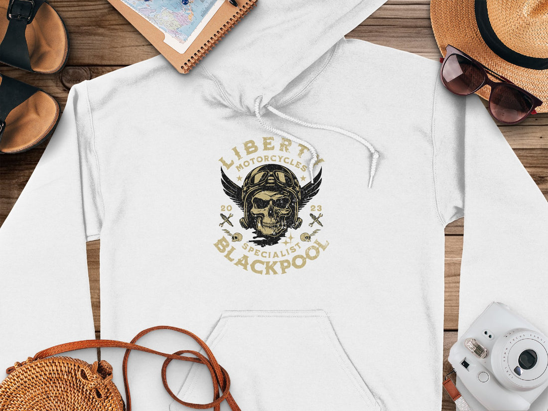 A white hoodie featuring a winged skull and Liberty Motorcycles Specialist Blackpool 2023 by Garment Graphics sits on wood, surrounded by a hat, sunglasses, sandals, camera, notebook, and woven bag—ideal companions for the iconic Blackpool hoodies style.