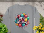 A gray unisex sweatshirt by Garment Graphics is on display against a textured background, featuring vibrant balloons and Happy Birthday in a playful font. Ideal for celebrations, its complemented with green and yellow plants nearby.