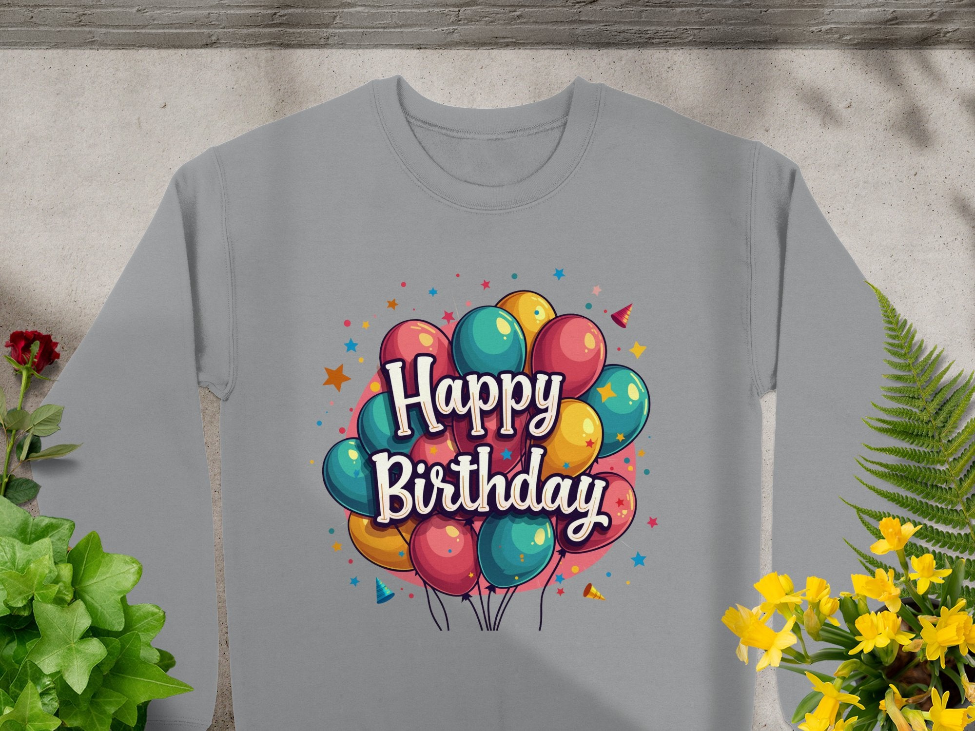 A gray unisex sweatshirt by Garment Graphics is on display against a textured background, featuring vibrant balloons and Happy Birthday in a playful font. Ideal for celebrations, its complemented with green and yellow plants nearby.