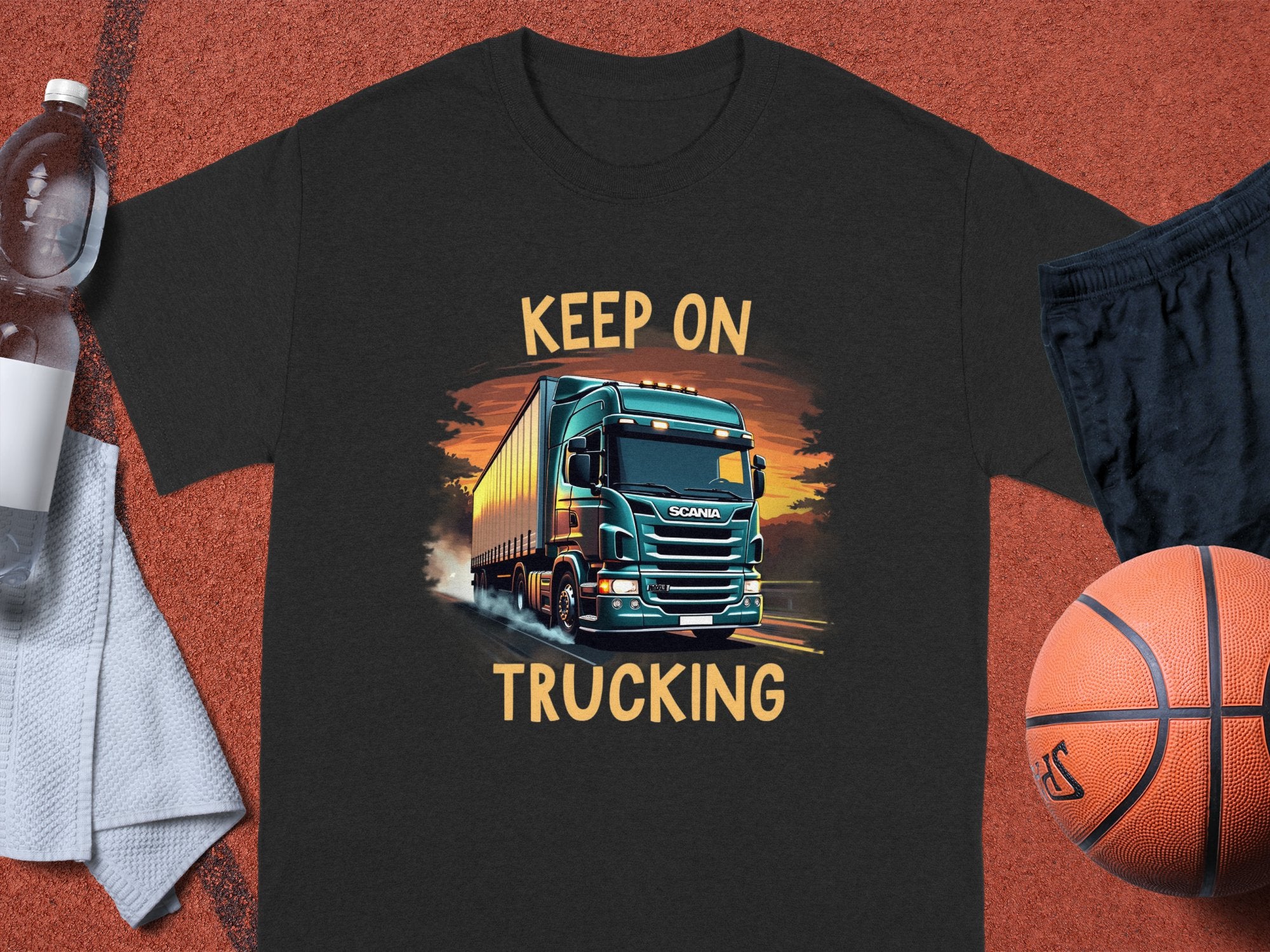 A black Garment Graphics t-shirt displays a large green truck driving into the sunset, with Keep On Trucking above and below. Perfect for truck enthusiasts, its styled with a water bottle, towel, shorts, and basketball nearby.