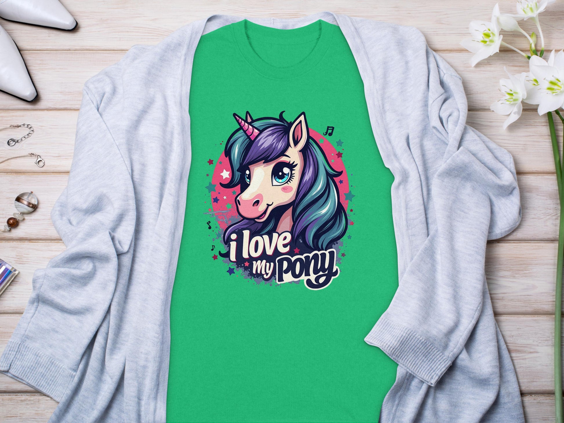 A Garment Graphics green unicorn enthusiast T-shirt with a cartoon unicorn and I love my pony text is displayed on wood alongside a light grey cardigan, white flowers, high-heeled shoes, and accessories like a bracelet and rings.