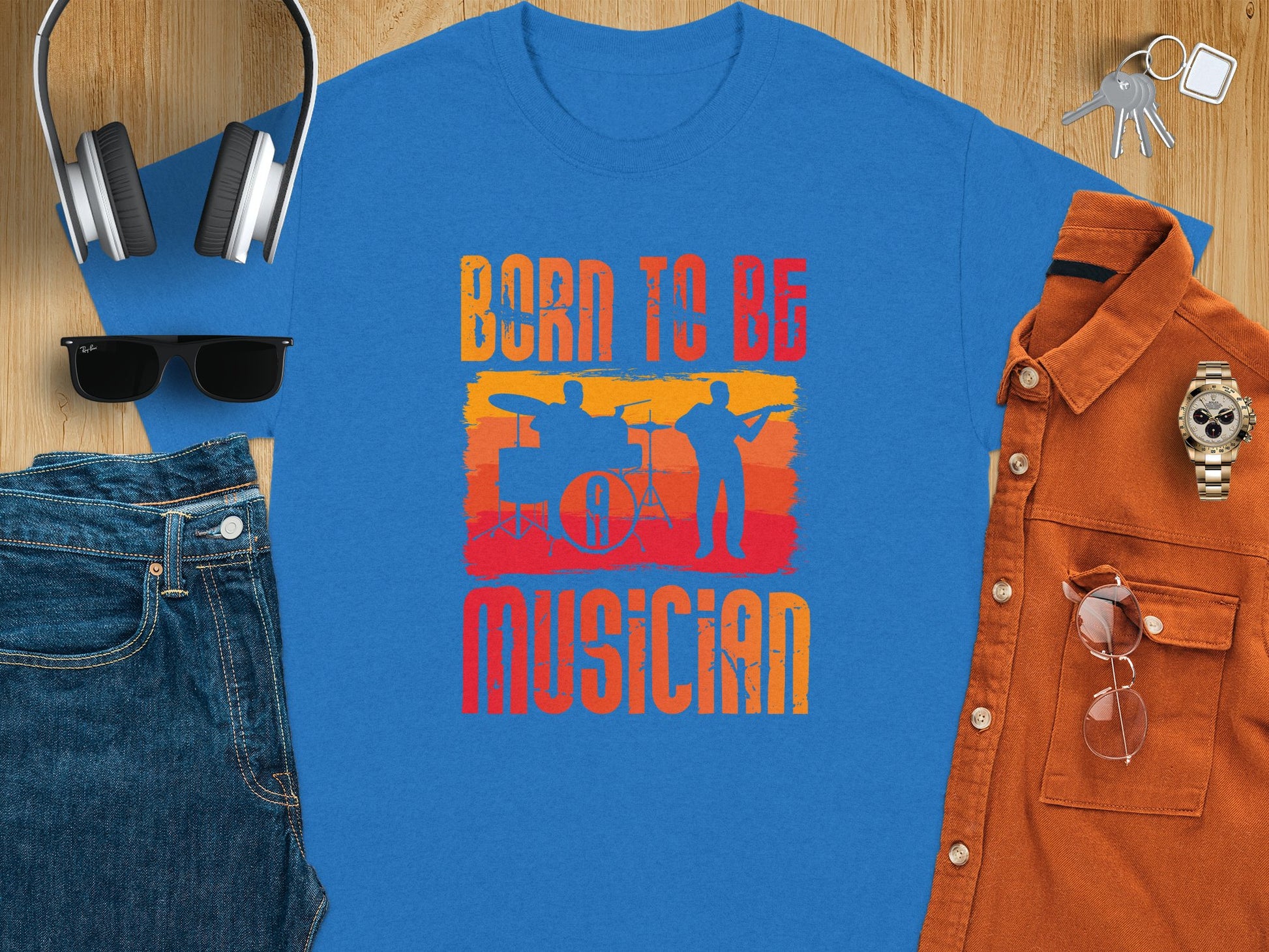 The vibrant orange and yellow Born To Be Musician T-shirt by Garment Graphics features silhouettes of a drummer and guitarist. Ideal for music lovers, its styled with blue jeans, sunglasses, a blue cap, headphones, a brown shirt, keys, and a wristwatch on a wood surface.
