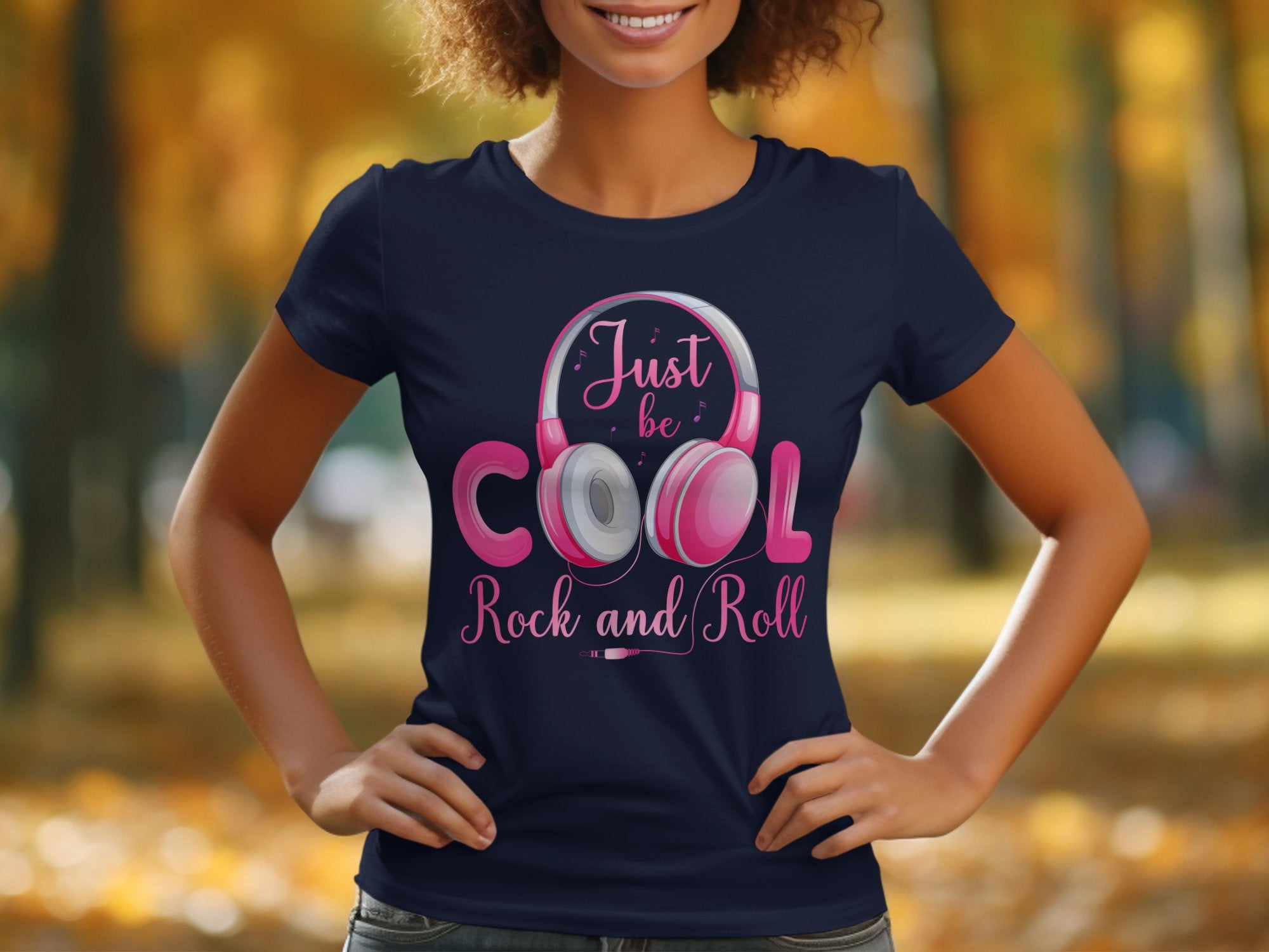 A person wears a Garment Graphics navy blue t-shirt with pink headphones and Just be cool, Rock and Roll text, set against a blurred autumn backdrop of trees and fallen leaves—ideal for music lovers who enjoy stylish headphone-themed attire.