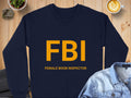 This Garment Graphics sweatshirt is navy blue with FBI in bold yellow and FEMALE BOOB INSPECTOR beneath. It offers unisex sizing and is showcased on a wooden surface with a jacket, plant, and latte.