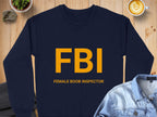 This Garment Graphics sweatshirt is navy blue with FBI in bold yellow and FEMALE BOOB INSPECTOR beneath. It offers unisex sizing and is showcased on a wooden surface with a jacket, plant, and latte.