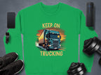 A green Garment Graphics sweatshirt for truck enthusiasts features a truck illustration with KEEP ON TRUCKING. Nearby are items like a smartphone, smartwatch, wireless speaker, sneakers, and adjustable dumbbells on a gray surface.