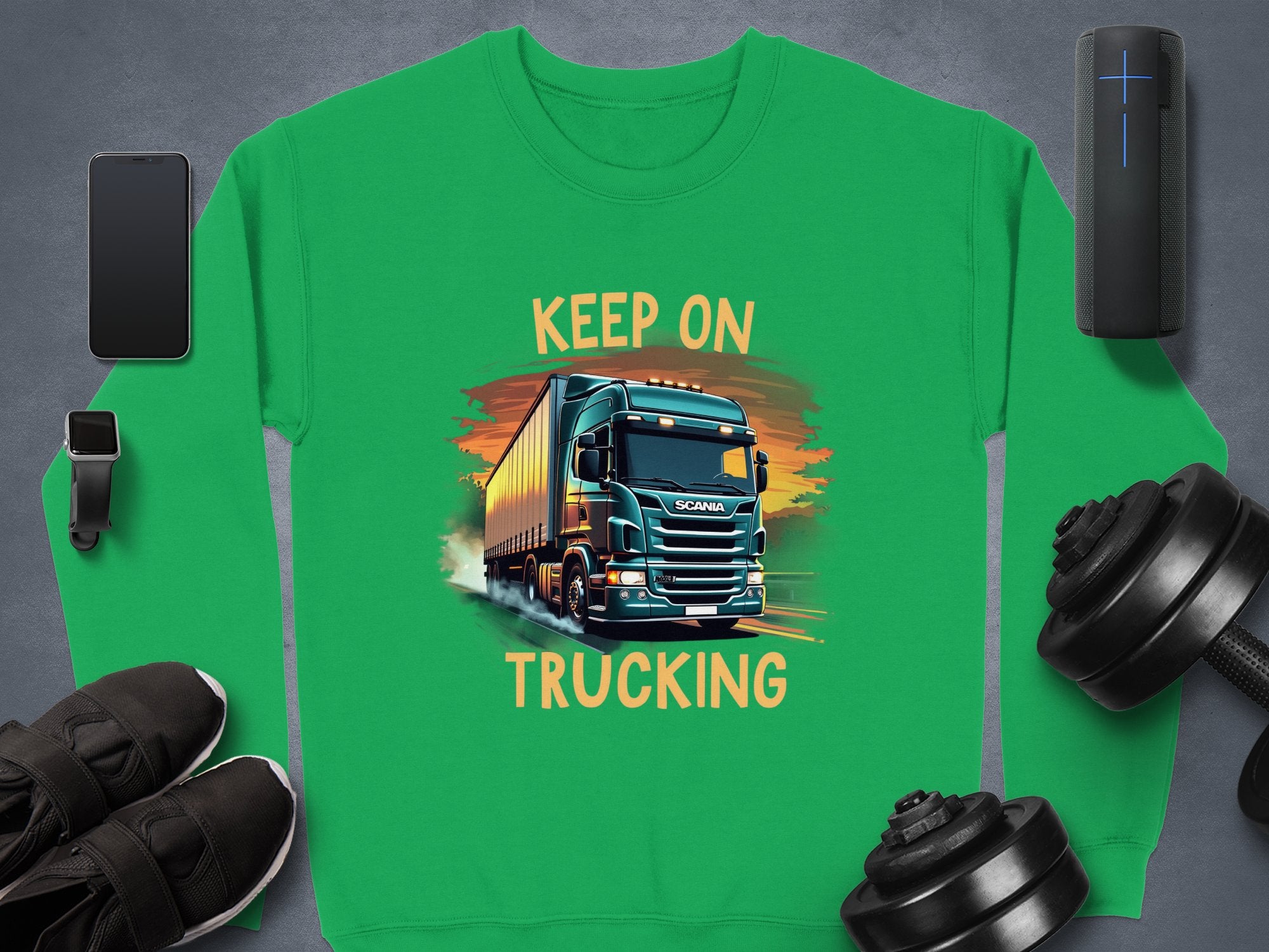 A green Garment Graphics sweatshirt for truck enthusiasts features a truck illustration with KEEP ON TRUCKING. Nearby are items like a smartphone, smartwatch, wireless speaker, sneakers, and adjustable dumbbells on a gray surface.