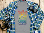 A Garment Graphics gray T-shirt featuring a vibrant Woodstock 1969 design exudes retro vibes on a wooden surface alongside a blue plaid shirt, headphones, camera, and white takeaway coffee cup, encapsulating the vintage festival era.