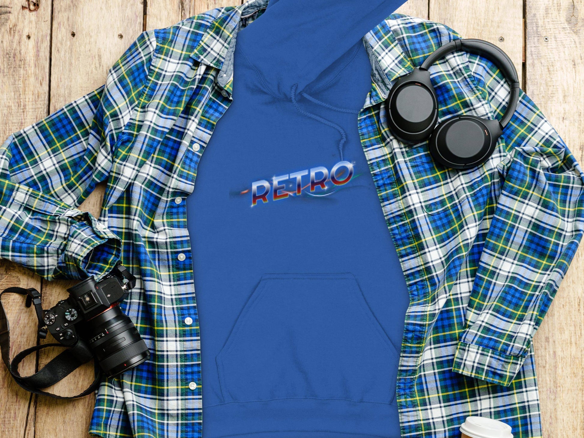 A vivid blue hoodie with Retro in stylized letters from Garment Graphics is laid flat on a wooden surface, surrounded by a plaid shirt, headphones, a camera, and a lidded coffee cup for a trendy modern vibe.