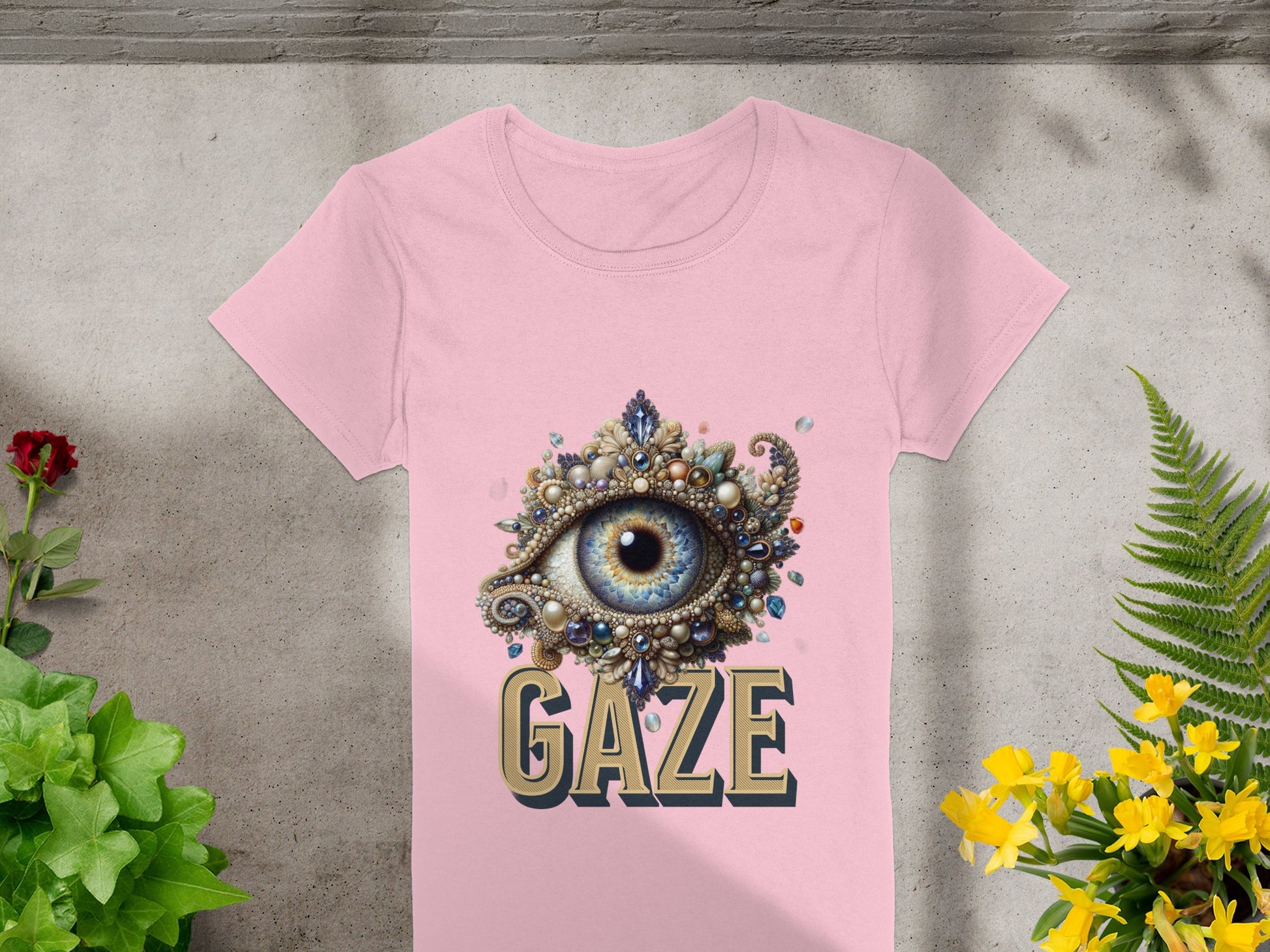 The Gaze graphic T-Shirt by Garment Graphics features a striking beaded eye design and the word GAZE. Photographed on concrete, its framed by green foliage and yellow flowers, and tailored for women.