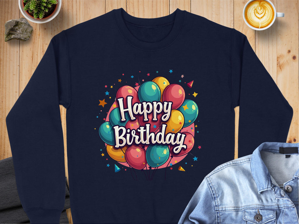 A navy Happy Birthday sweatshirt by Garment Graphics, featuring colorful balloons, is perfect for celebrations. Casually set on a wooden surface beside a denim jacket, cappuccino cup, plant, and stones, its unisex sizing makes it ideal for spreading birthday cheer to anyone.
