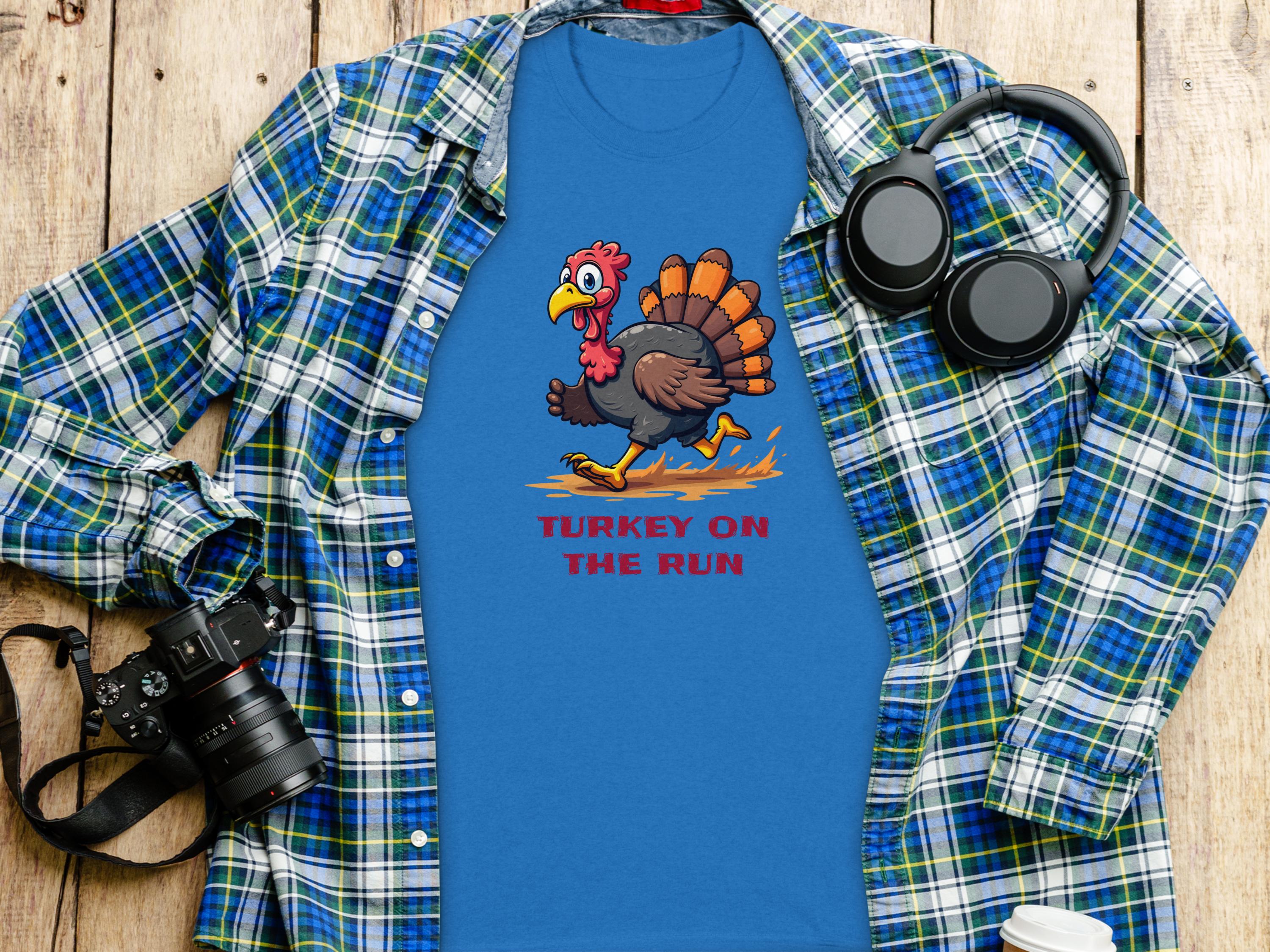A Garment Graphics blue T-shirt, adorned with a whimsical cartoon turkey and the caption Turkey on the Run, is displayed on a wooden surface, accompanied by a blue plaid shirt, headphones, a camera, and a takeaway coffee cup. Ideal for fans of humorous graphic tees.