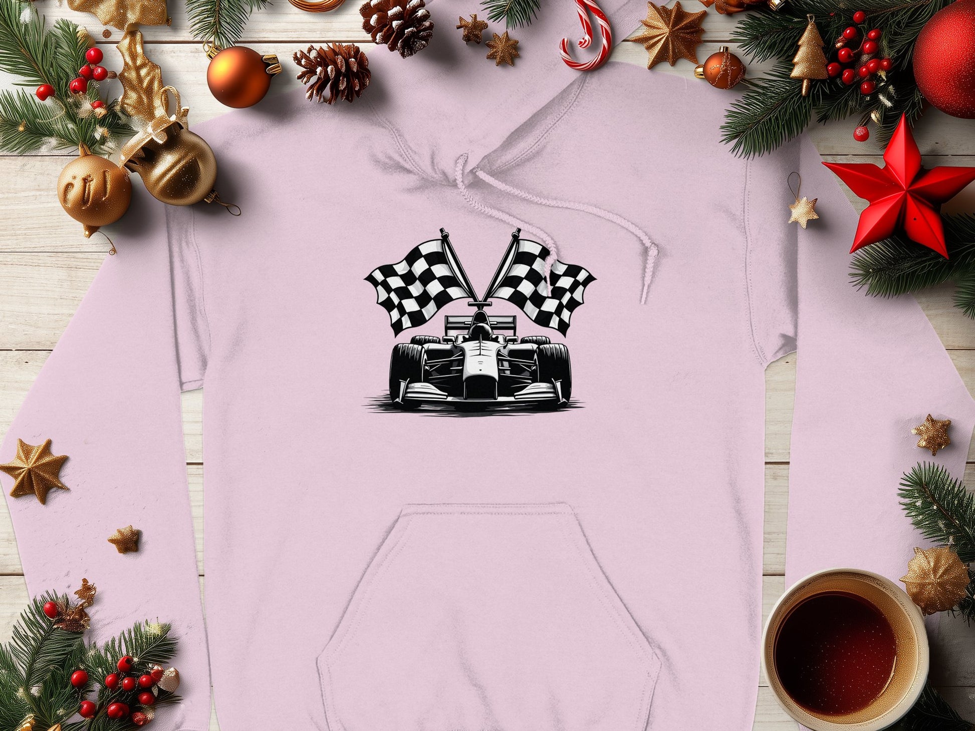 A Garment Graphics light pink hoodie with a racing car and crossed checkered flags design is laid flat, ideal for motorsport fans. It’s surrounded by festive decorations like pinecones, ornaments, and a warm drink on a wooden surface.