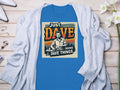 A blue Just Dave Doing Dave Things graphic T-shirt by Garment Graphics is displayed on a wooden surface with a classic fit, accompanied by a light gray cardigan, flowers, and scattered jewelry such as rings and a bangle.